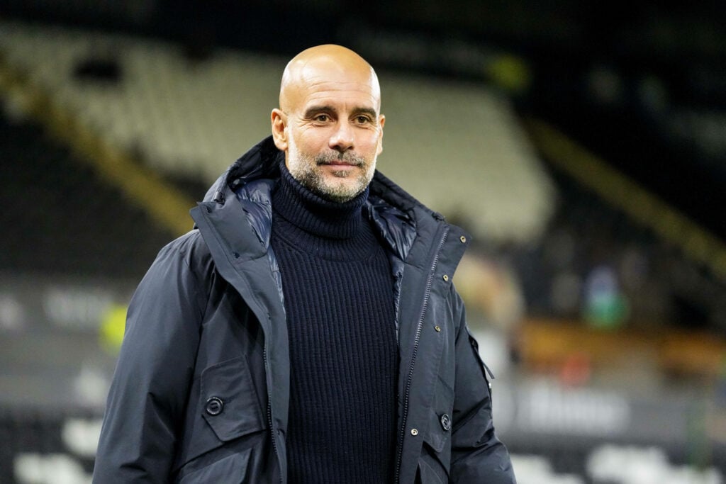 Manchester City manager Pep Guardiola smiles while standing on the touchline.