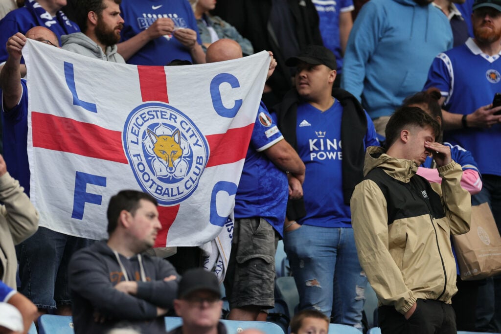 Leicester City fans hold a flag aloft as they look frustrated in the stands.