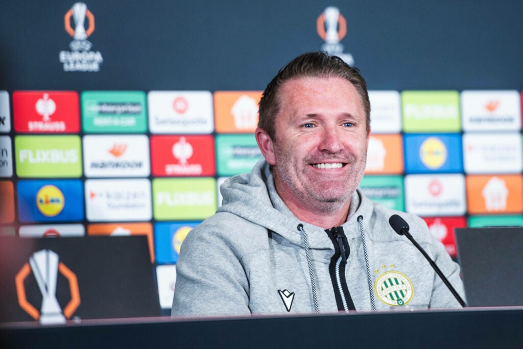 Ferencvaros manager Robbie Keane grins while sitting in a press conference.